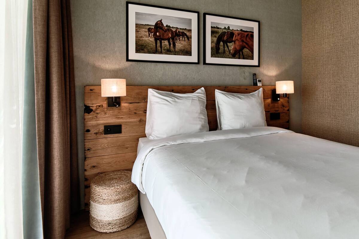 A hotel room with a wooden headboard, two framed horse pictures, a bed with white linens, two bedside lamps, and a woven basket.