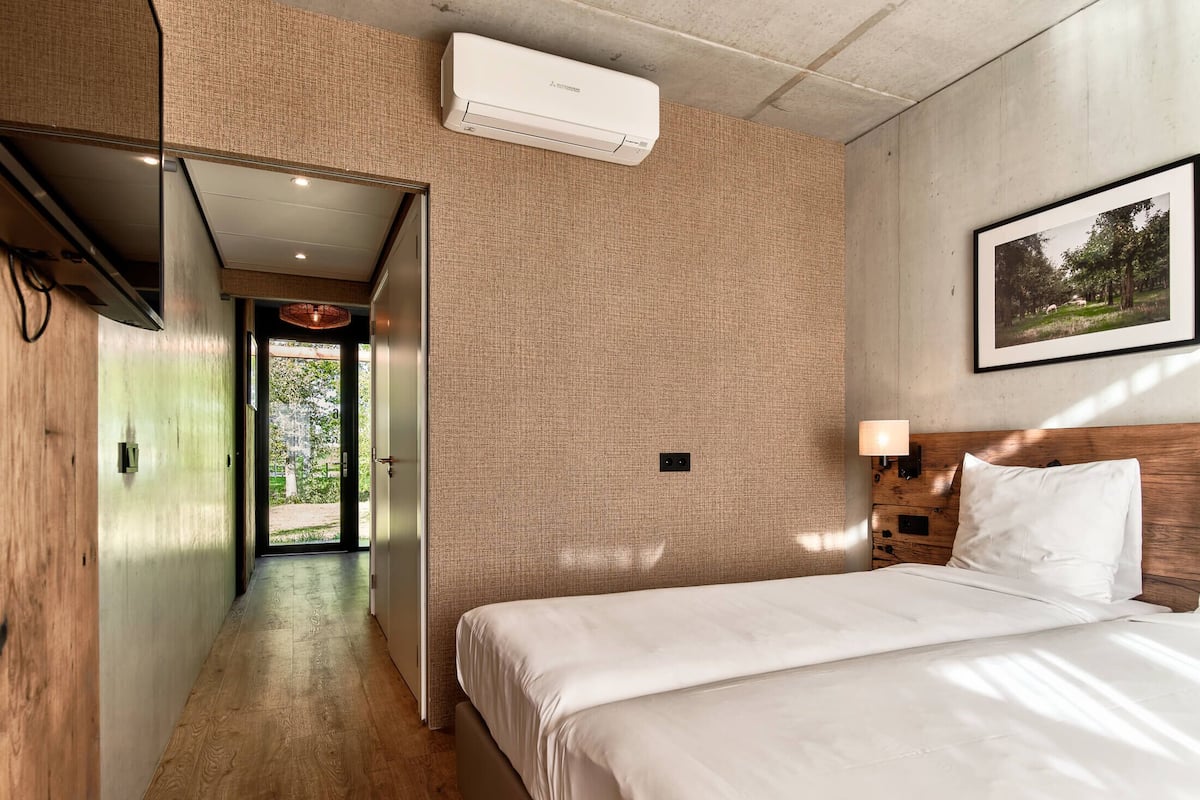 A hotel room with a wooden floor, a bed with white bedding, a mounted air conditioner, a framed picture on the wall, and a door leading to a corridor.