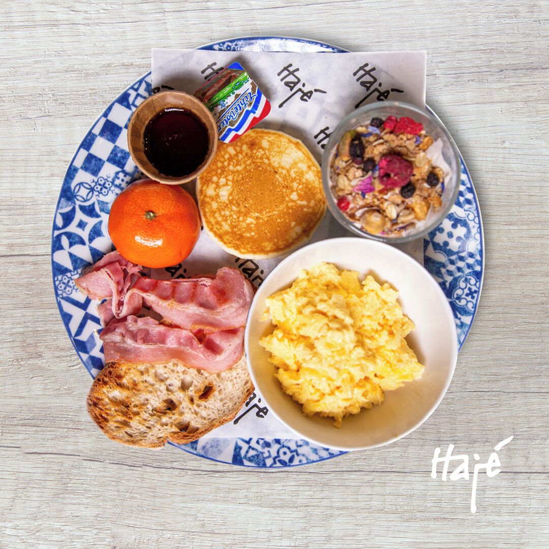 A breakfast plate with pancakes, scrambled eggs, bacon, toast, orange, and a bowl of cereal.