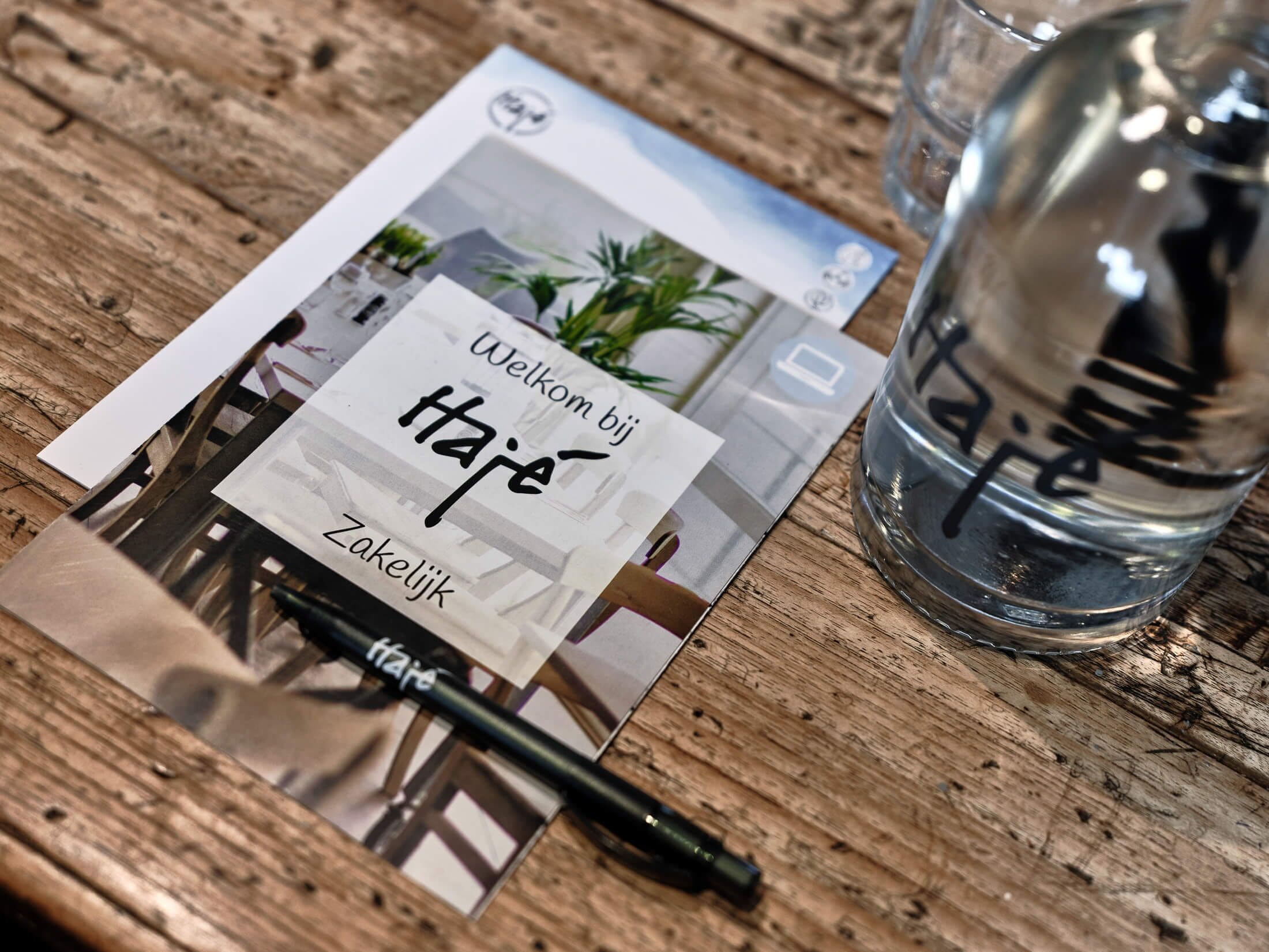 A wooden table with a glass of water, a pen, and a welcome card with a photo and text.
