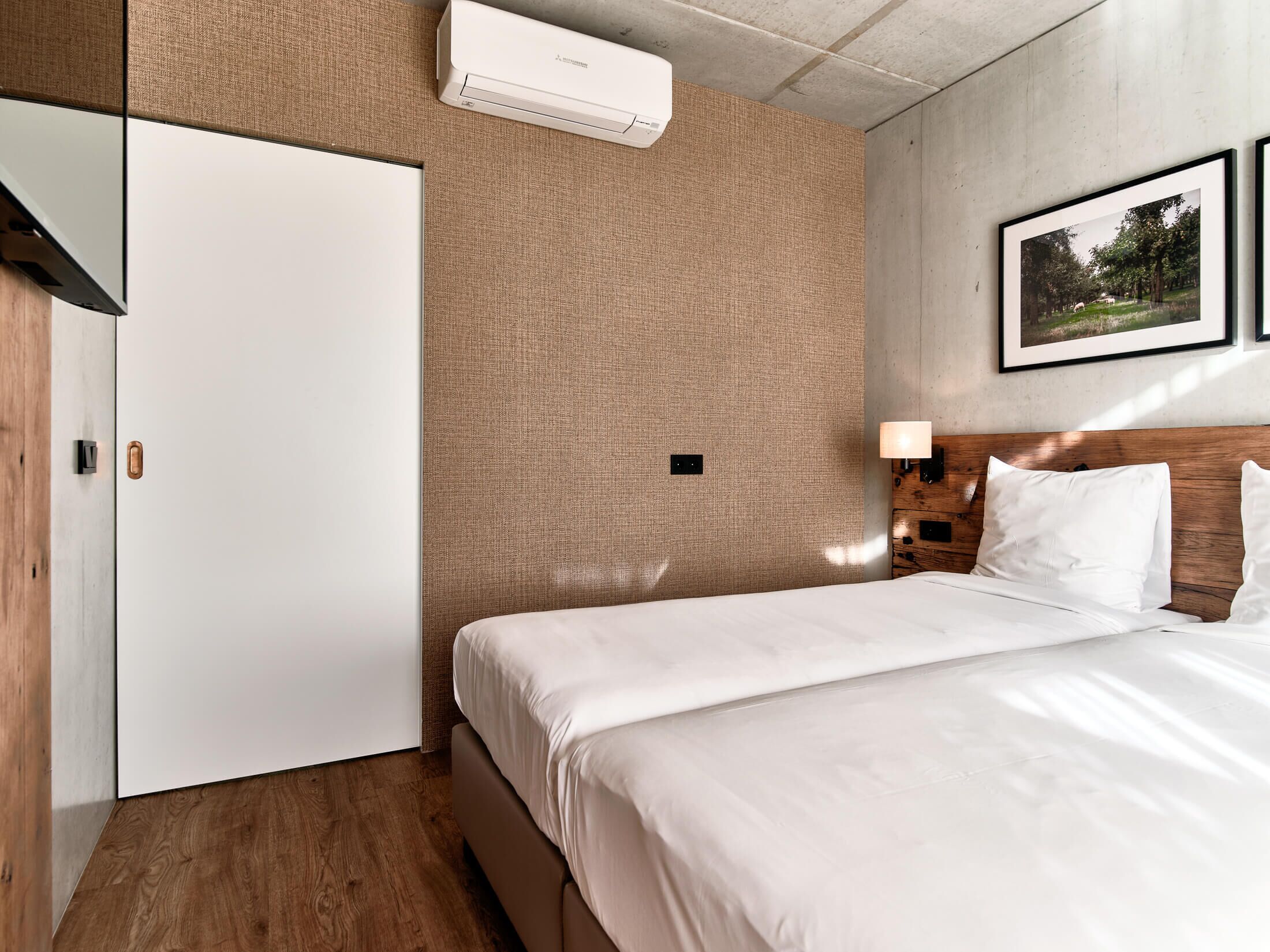 A hotel room with a bed, a wardrobe, an air conditioning unit, a wall-mounted lamp, and a framed picture on the wall.