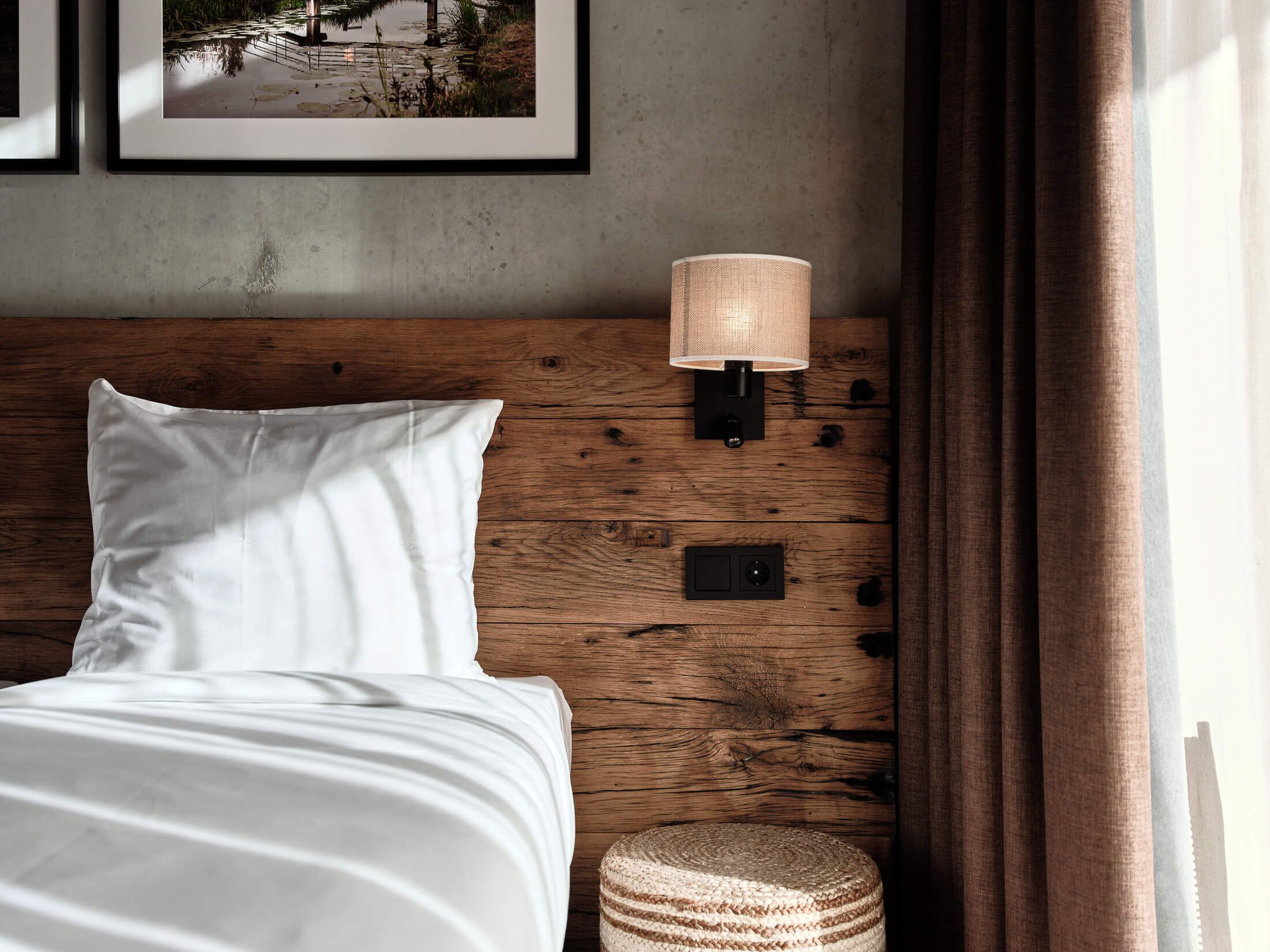 Una cama bien tendida con sábanas blancas, cabecera de madera, lámpara de pared y un pequeño taburete tejido.