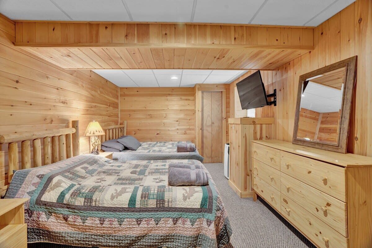 A wooden cabin room with two beds, a dresser, a TV, and a door leading outside.