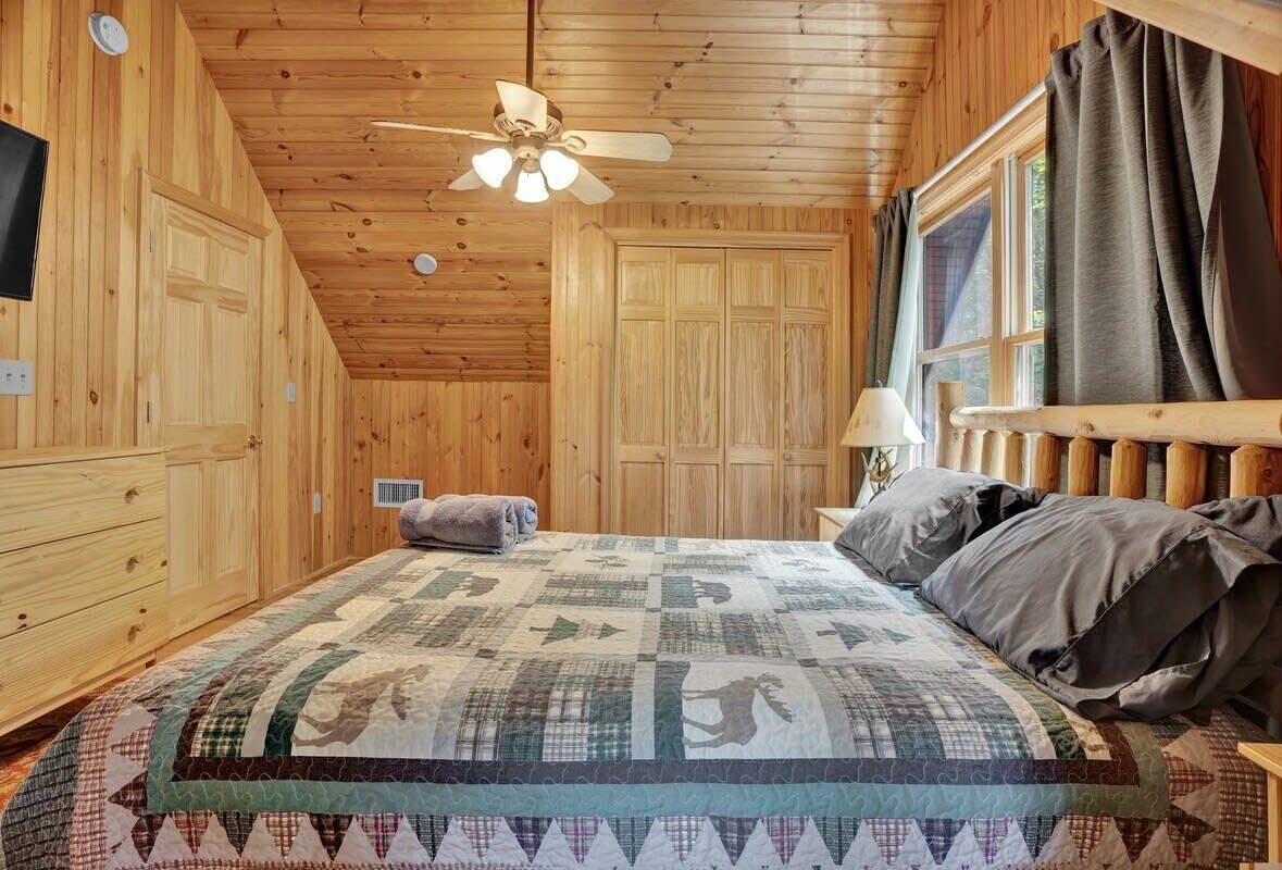 Een slaapkamer met een houten plafond, een bed met een sprei, een plafondventilator en een raam met gordijnen.