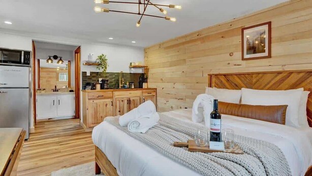 A modern hotel room with a wooden headboard, a bed with white linens, a bedside table with a bottle and glasses, and a view of the kitchen and bathroom through an open door. - Elements Pocono Cabin | Pickleball | Firepits (Barrett Township)
