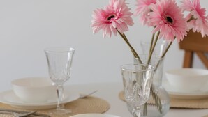 Ein Esstisch mit weißen Tellern, klarem Glas und rosafarbenen Blumen in einer Vase.