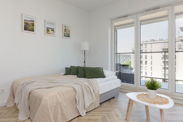 A modern bedroom with a bed, a bedside table with a plant, and a view of buildings through large windows. - Sunny Studio in Warsaw | Balcony and Private Parking (Warszawa)