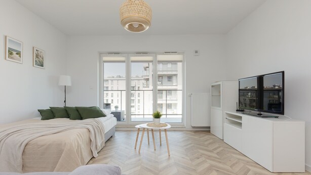 Ein moderner, gut beleuchteter Raum mit einem Bett, einem kleinen runden Tisch mit Pflanze, einem Fernseher auf einem weißen Schrank und großen Fenstern mit Blick auf die Stadt.