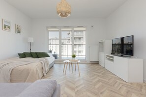 Ein moderner, gut beleuchteter Raum mit einem Bett, einem kleinen runden Tisch mit Pflanze, einem Fernseher auf einem weißen Schrank und großen Fenstern mit Blick auf die Stadt.
