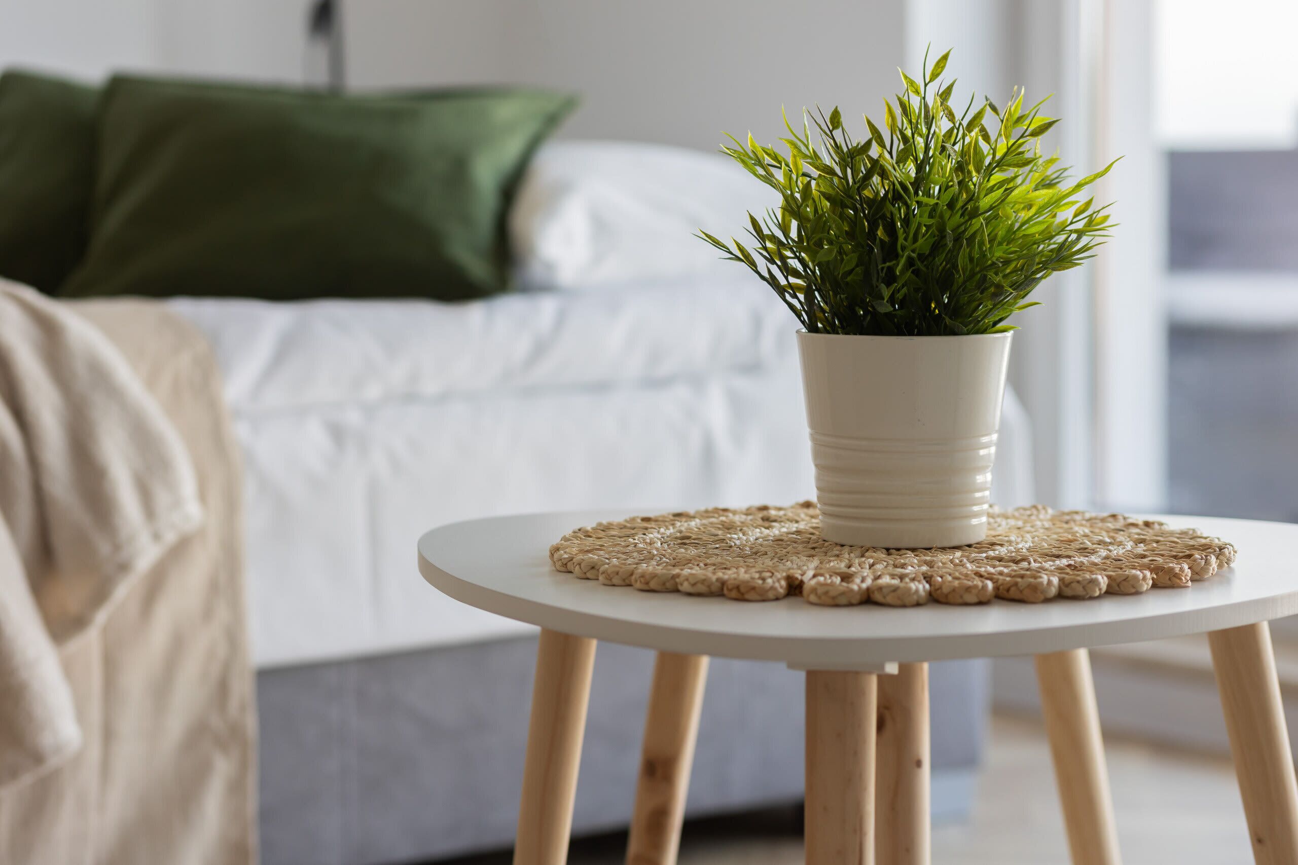 A potted plant on a round table with a woven placemat, a bed with white bedding and a green pillow in the background.