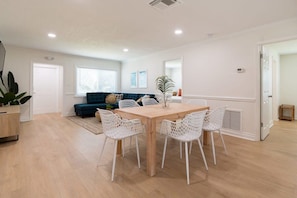 A modern living room with a wooden dining table, white chairs, a blue sofa, and a large window. - Tropical 7-unit retreat w/ pool & BBQ (Cocoa Beach)