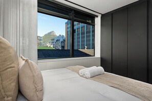 Una camera d'albergo con un letto, una finestra che si affaccia su edifici e una collina, e una parete scura.