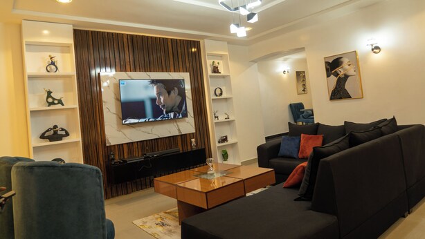 Een moderne woonkamer met een flatscreen televisie, een donkere bank, een houten salontafel en decoratieve planken.