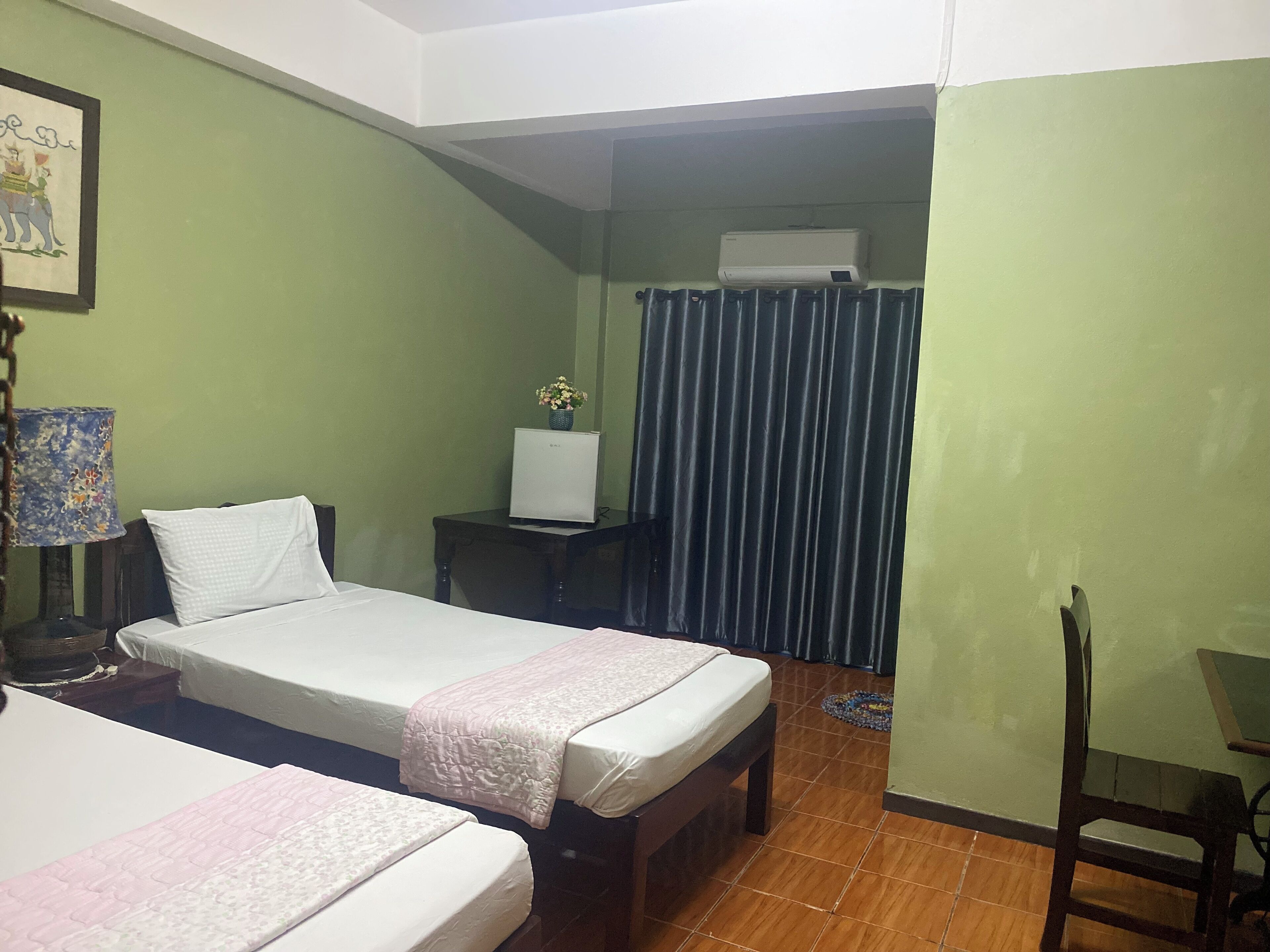 A hotel room with two beds, a small table, a chair, a lamp, a refrigerator, and a curtain.