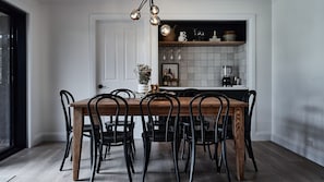 Uma área de refeições com mesa de madeira e cadeiras pretas, uma porta branca e uma cozinha com azulejos na parede e diversos utensílios.