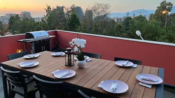 Ein Dachterrassen-Essbereich mit einem Holztisch für vier Personen, einem Grill und Blick auf die Skyline bei Sonnenuntergang.