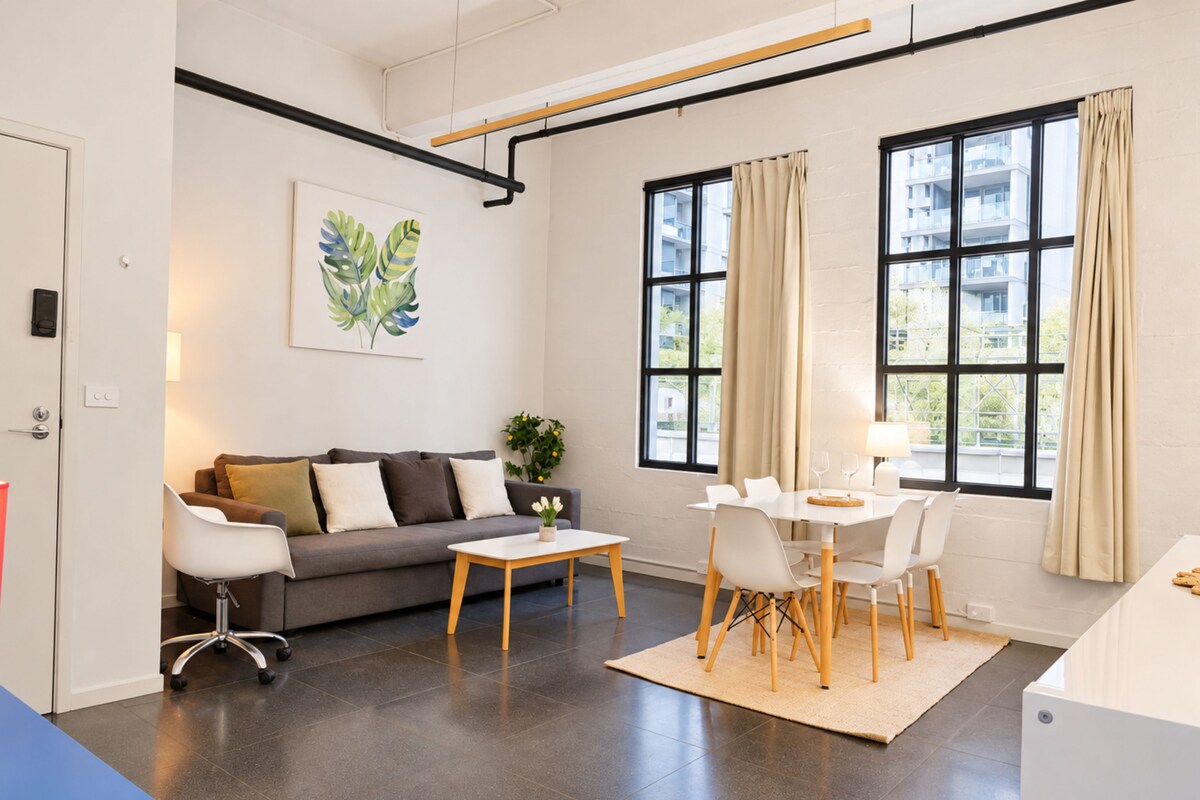 A modern living room with a sofa, coffee table, dining table, and chairs. There is a large window with curtains, a plant on the coffee table, and a painting on the wall.
