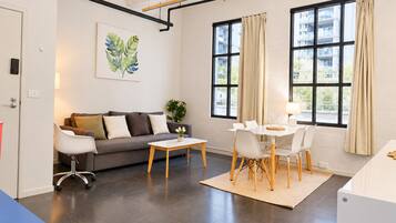 A modern living room with a sofa, coffee table, dining table, and chairs. There is a large window with curtains, a plant on the coffee table, and a painting on the wall.