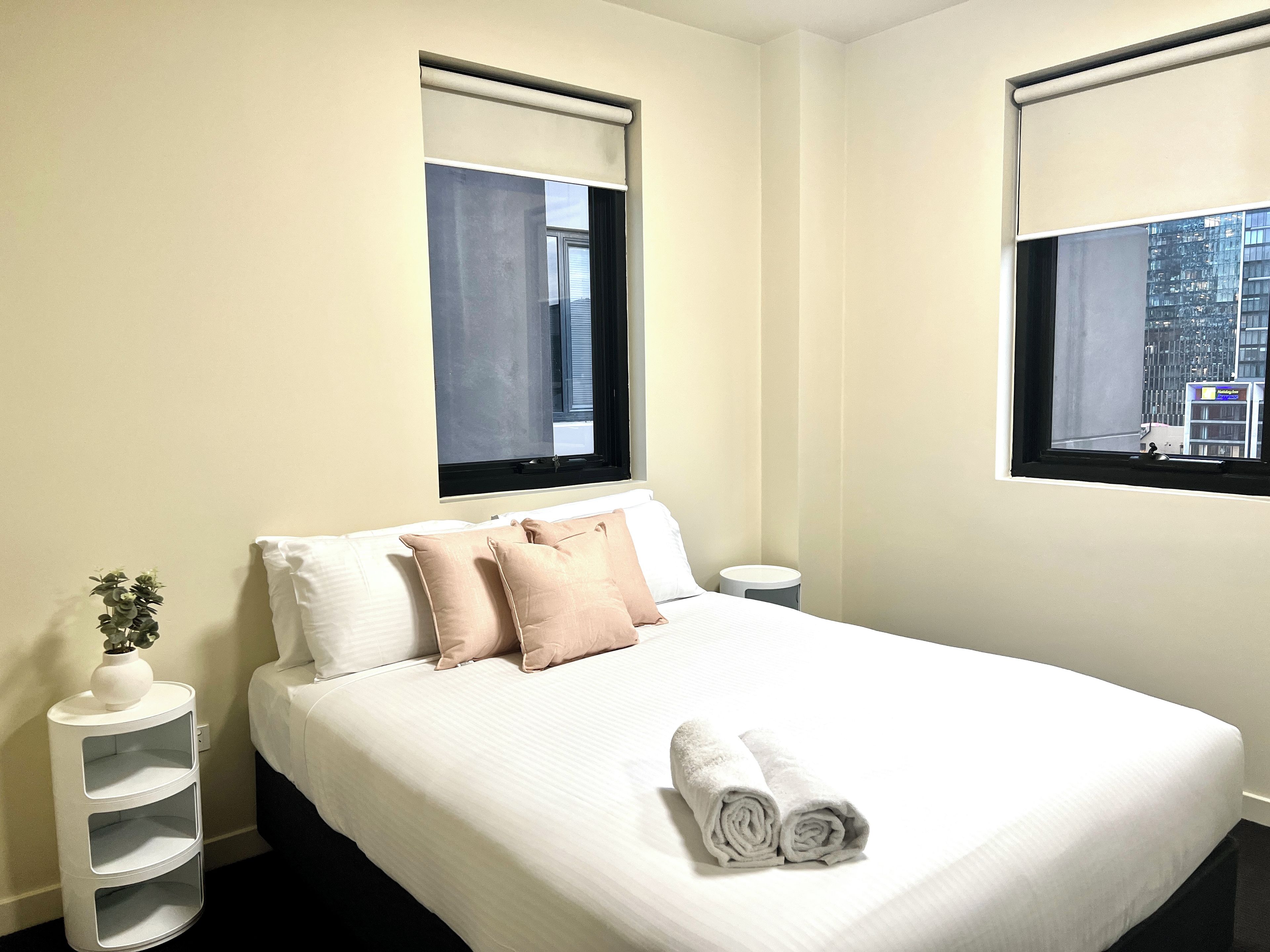 A hotel room with a bed, two windows with blinds, a bedside table with a vase, and a view of a cityscape.