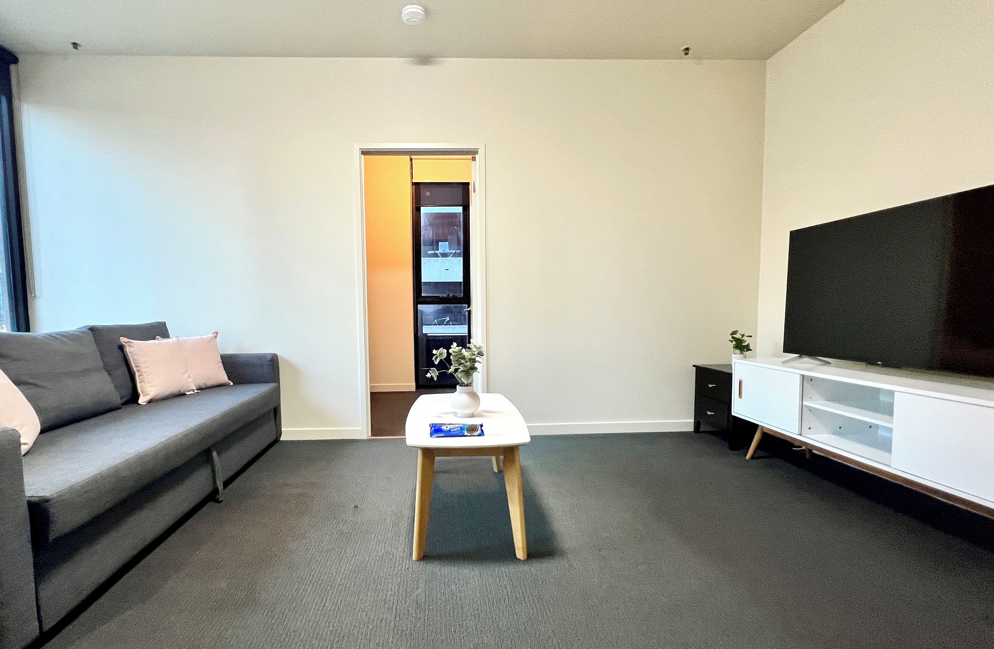 A living room with a grey sofa, a white coffee table with a plant and a snack, a TV on a white cabinet, and a door leading to a balcony.