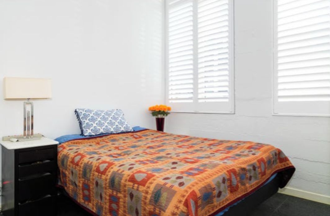 A bedroom with a bed, a nightstand, a lamp, a window with blinds, and a vase with flowers.