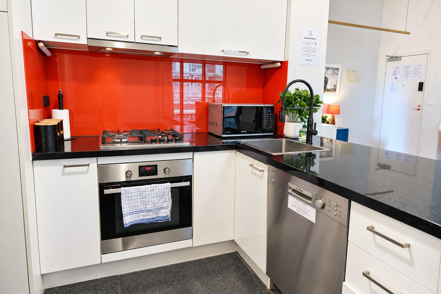 Una cocina moderna con armarios blancos, salpicadero rojo, electrodomésticos de acero inoxidable, incluyendo microondas, horno y lavavajillas, y encimera negra.