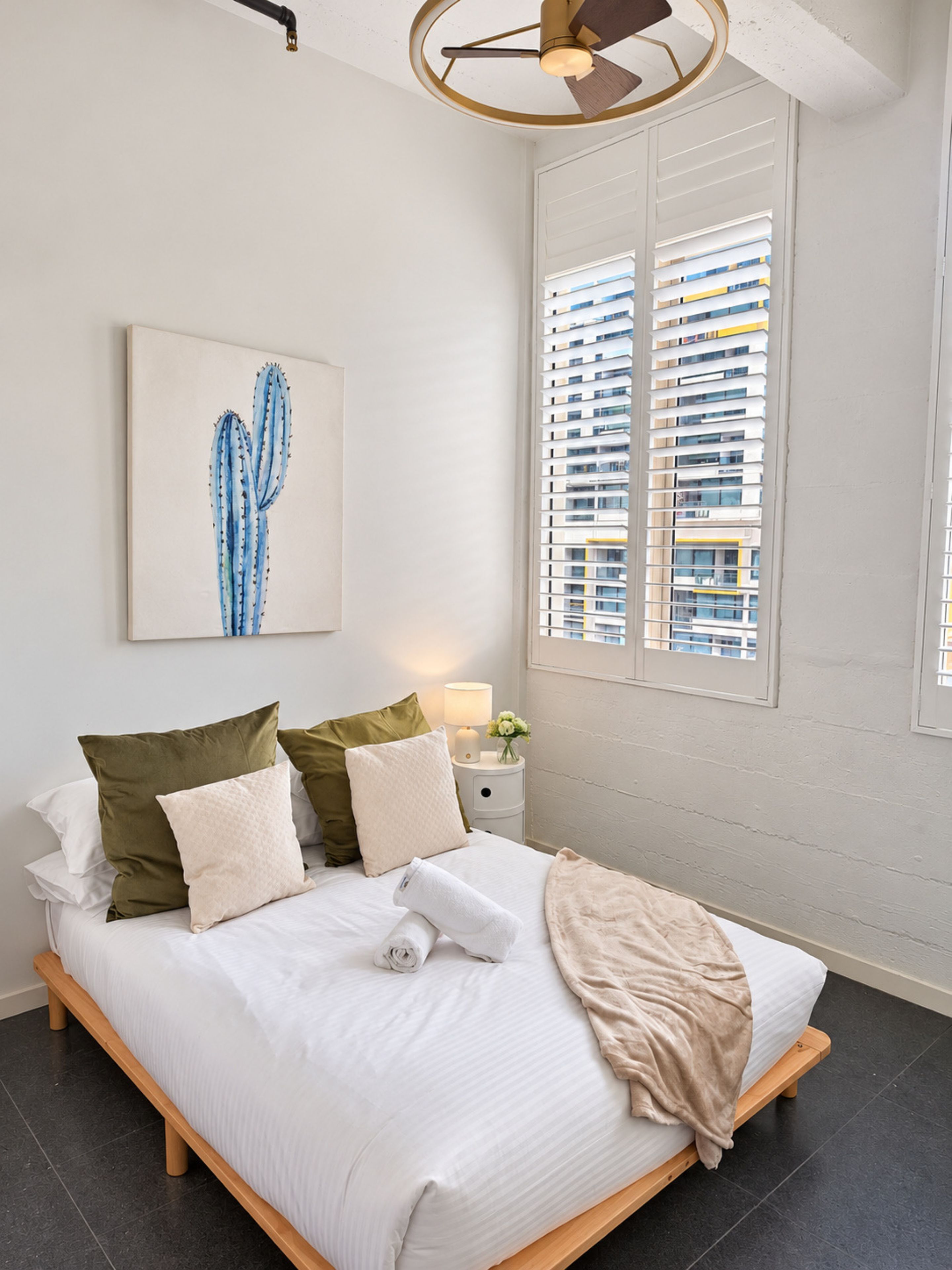 A modern bedroom with a bed, pillows, a bedside table with a lamp, a cactus artwork, and a window with blinds.