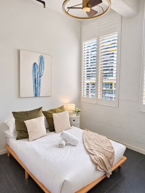 A modern bedroom with a bed, pillows, a bedside table with a lamp, a cactus artwork, and a window with blinds.