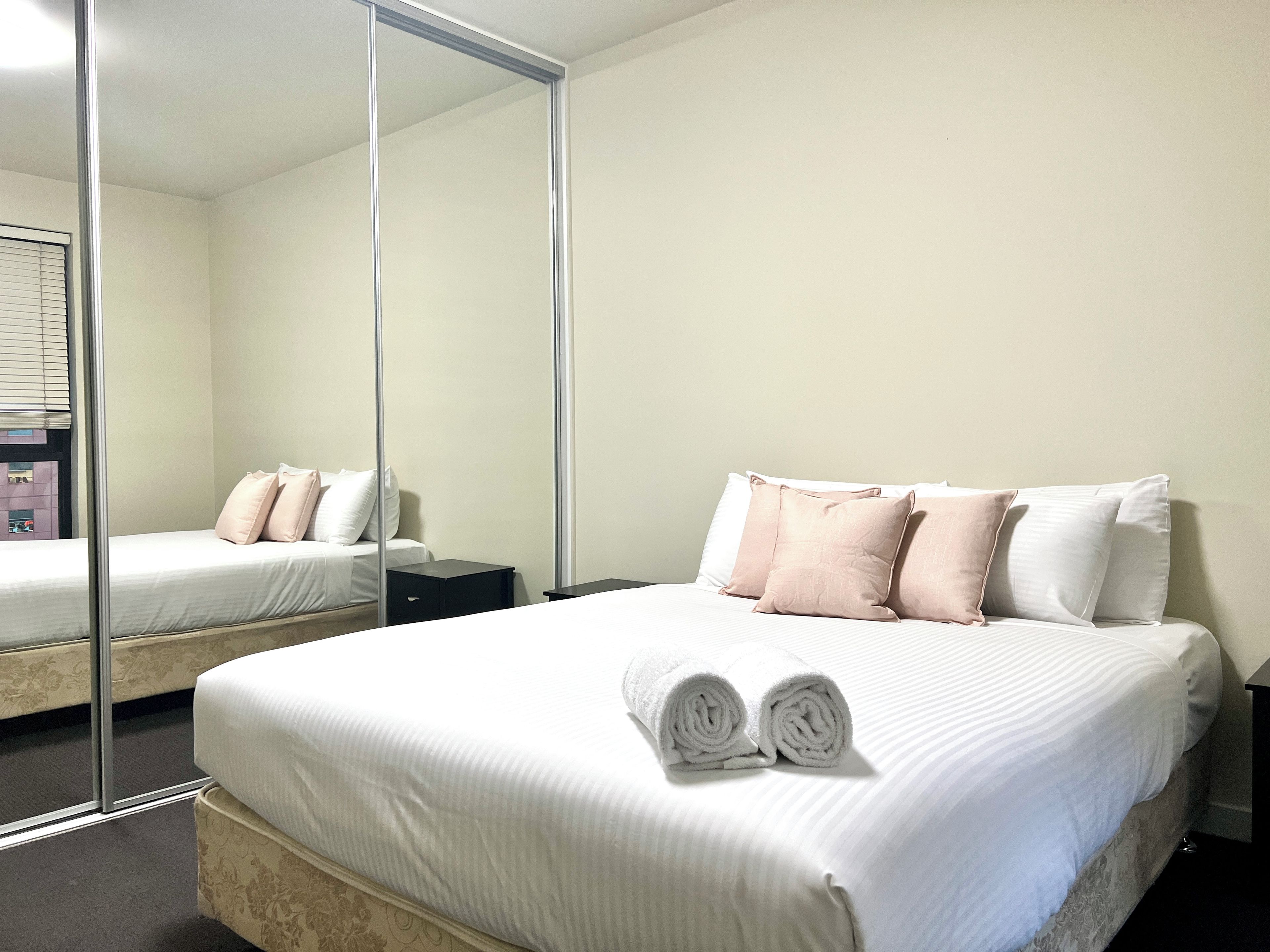 Une chambre d’hôtel avec un grand miroir, un lit avec une literie blanche et plusieurs oreillers, une petite table de chevet et une serviette roulée posée sur le lit.