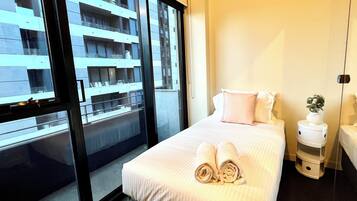 A hotel room with a large window, a bed with rolled towels, a bedside table, and a nightstand with a plant.