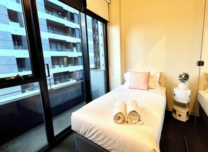 A hotel room with a large window, a bed with rolled towels, a bedside table, and a nightstand with a plant. - Readyset Apartments on Little Bourke (Melbourne)
