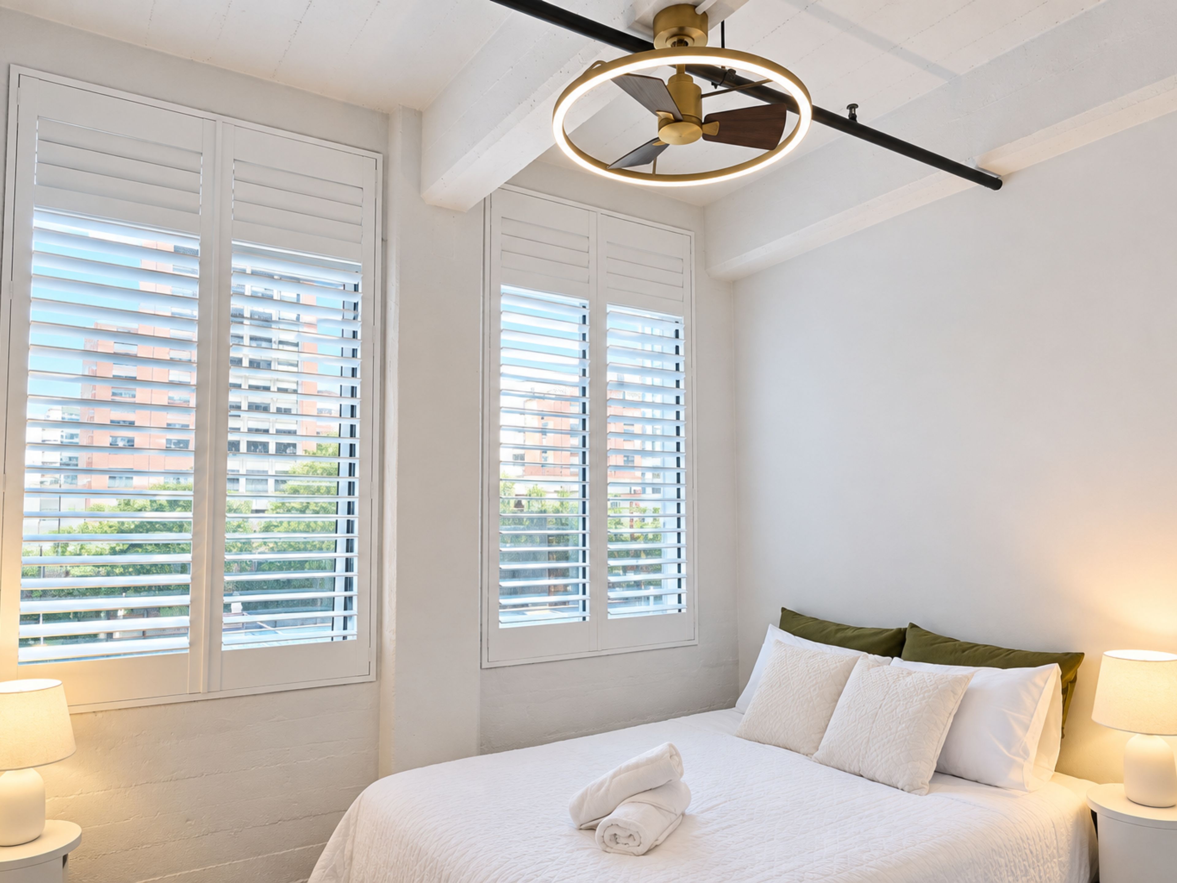 A hotel room with a bed, two bedside lamps, a ceiling fan, and large windows with blinds.