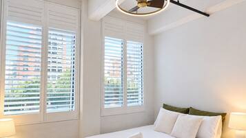 A hotel room with a bed, two bedside lamps, a ceiling fan, and large windows with blinds.