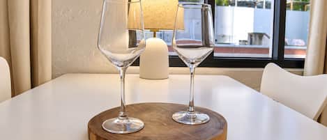 Una mesa con dos copas de vino sobre un posavasos de madera, una vela y una lámpara en una habitación con vistas a edificios a través de una ventana.
