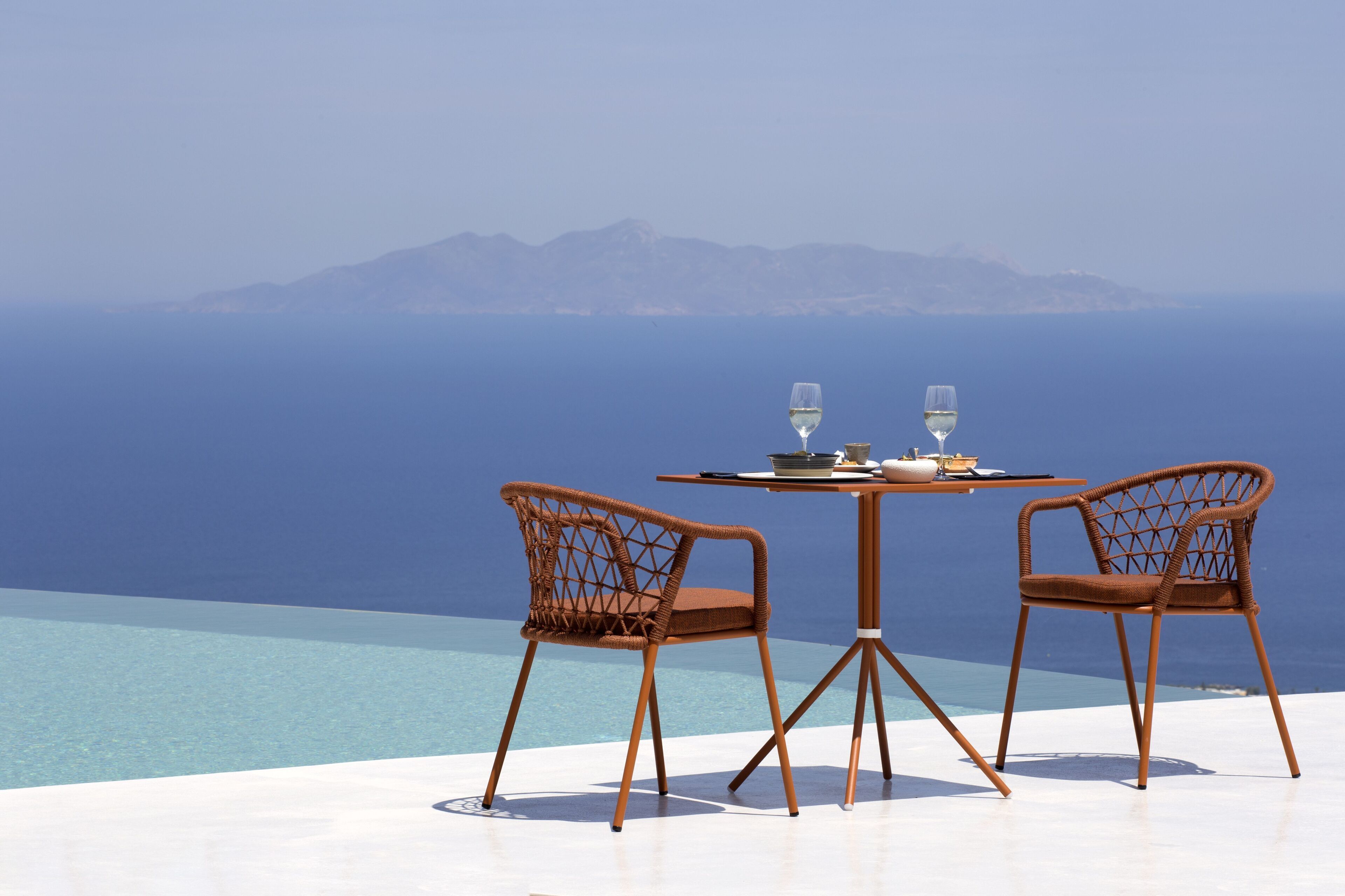 Una mesa con dos sillas y dos copas de vino en una terraza con vistas a un cuerpo de agua y montañas lejanas.