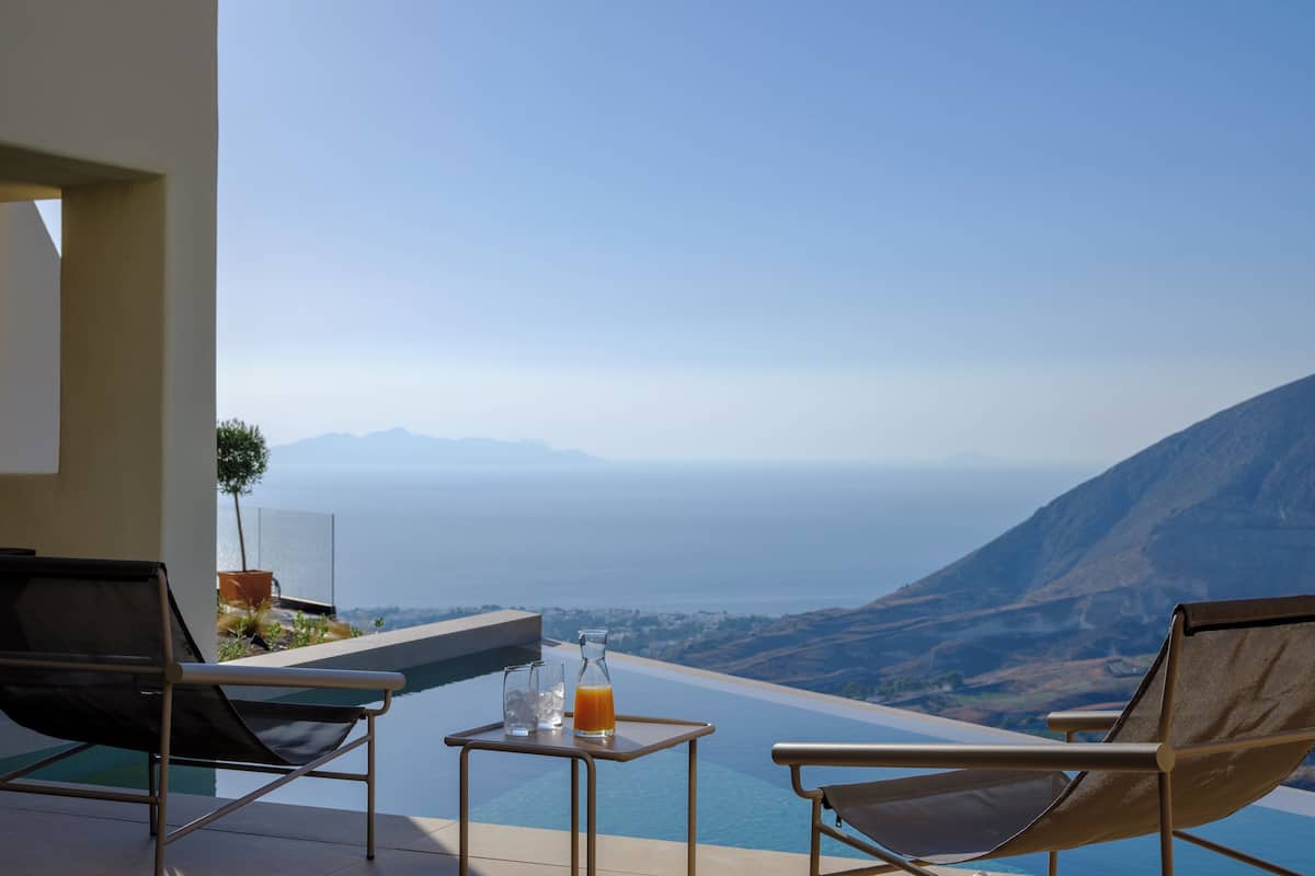 A poolside area with two lounge chairs and a table, overlooking a scenic view of the ocean and mountains.