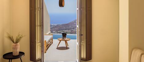 A room with a large sliding glass door leading to a balcony with a view of the sea and hills. There is a small round table with a bowl and a vase on it, and a sofa with a cushion.