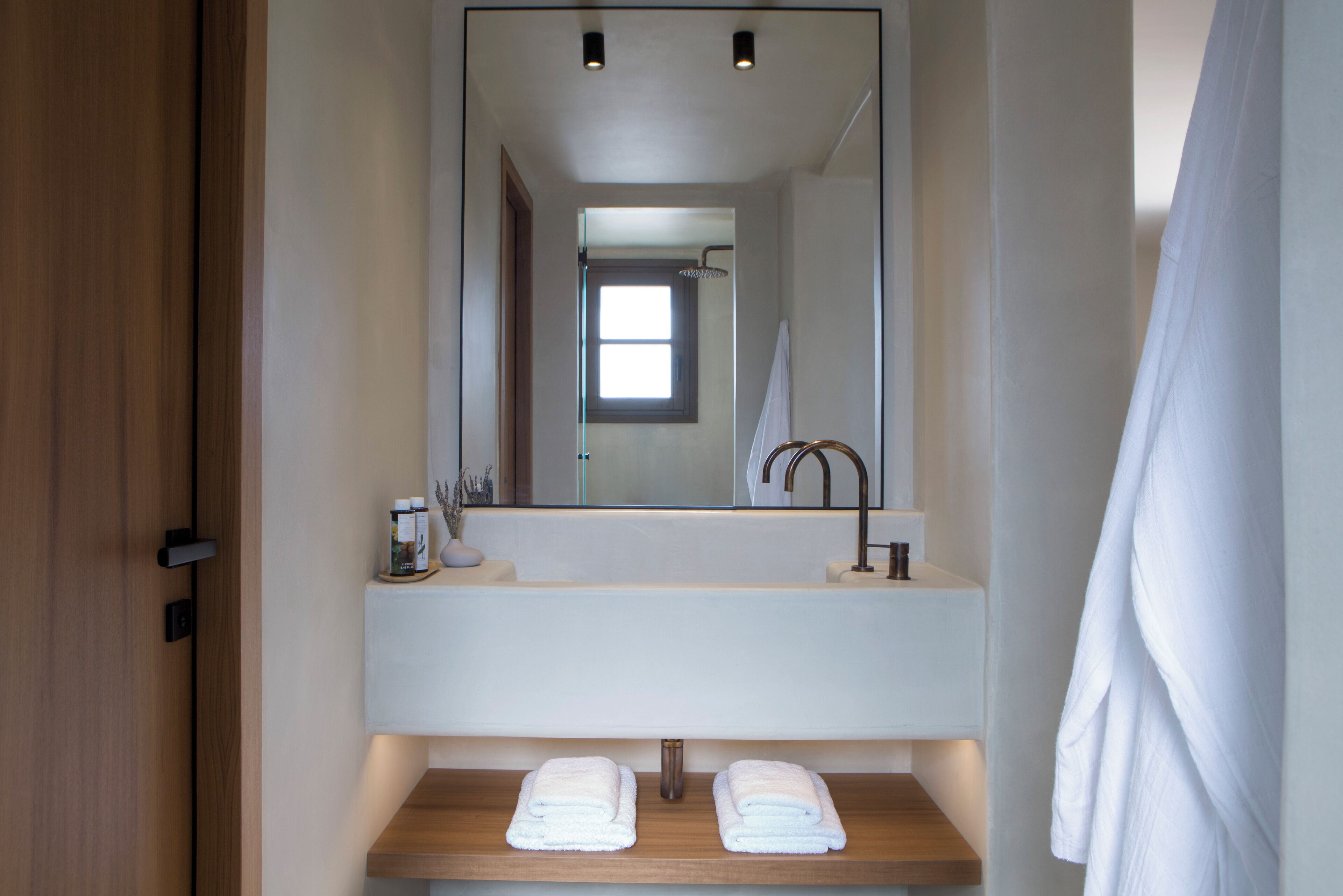 Une salle de bain moderne avec un meuble-lavabo en bois, un grand miroir et une zone de douche visible à travers l’ouverture de la porte.