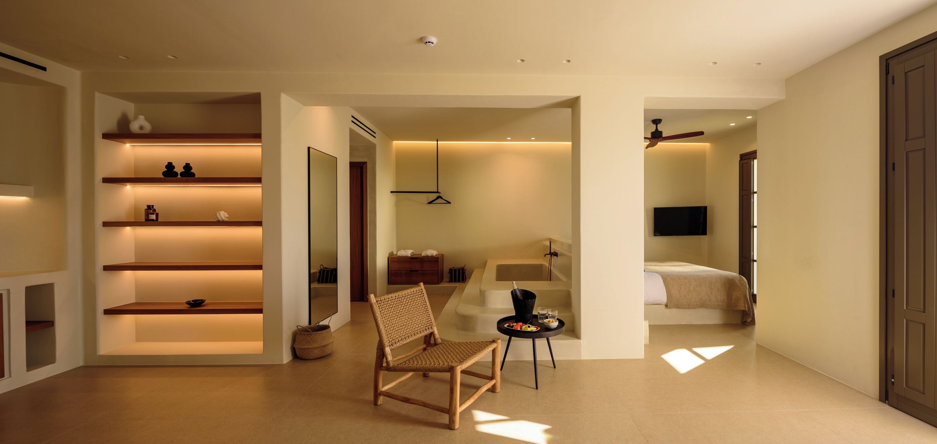 A modern hotel room with a wooden chair, a small table with a bowl of fruit, and a bed with a TV.