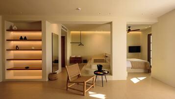 A modern hotel room with a wooden chair, a small table with a bowl of fruit, and a bed with a TV.