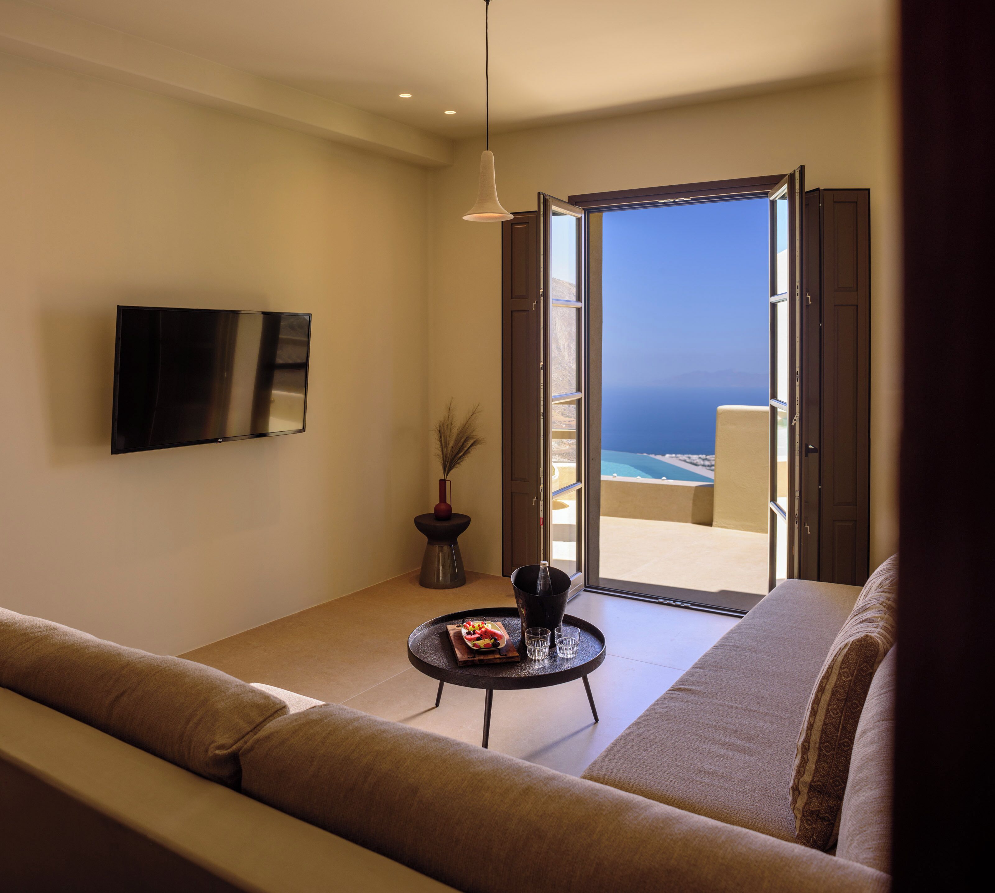 A modern living room with a sofa, a small table with a bottle and glasses, a TV mounted on the wall, and a view of the ocean through an open door.