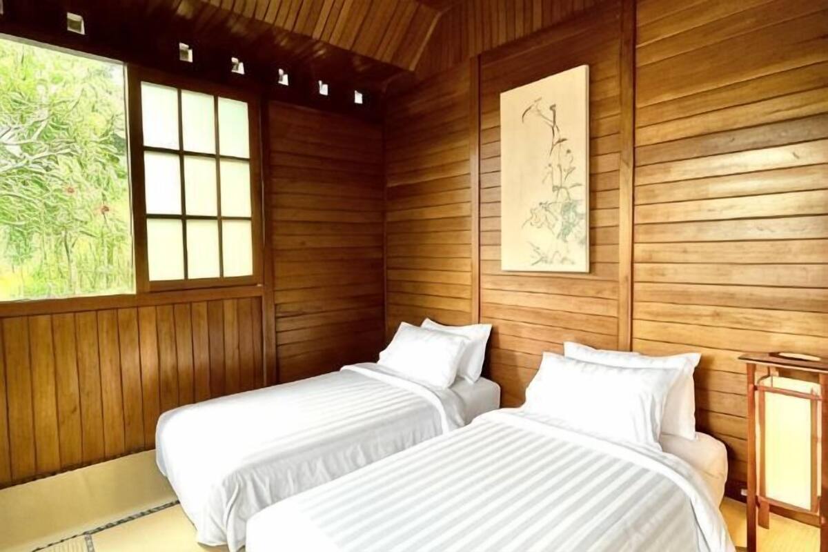 Quarto com paredes de madeira, duas camas com roupa de cama branca, uma janela com vista para o verde e uma obra de arte emoldurada na parede.
