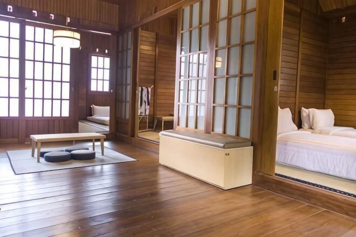 Quarto tradicional japonês com piso de tatame, cama de madeira baixa, um banco e uma porta de correr com painéis de vidro.
