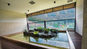 A tranquil indoor pool area with a wooden deck, a rock garden, and a view of a mountainous landscape through large windows. - The Onsen Hot Spring Resort (Batu)