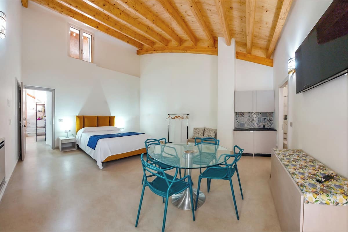 Una habitación con una cama, una mesa redonda de vidrio con sillas, un televisor y una kitchenette.