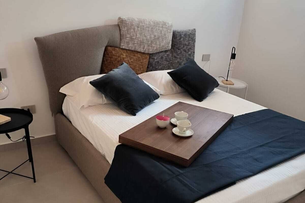 Una cama bien hecha con varios almohadones y una bandeja con tazas y tetera.