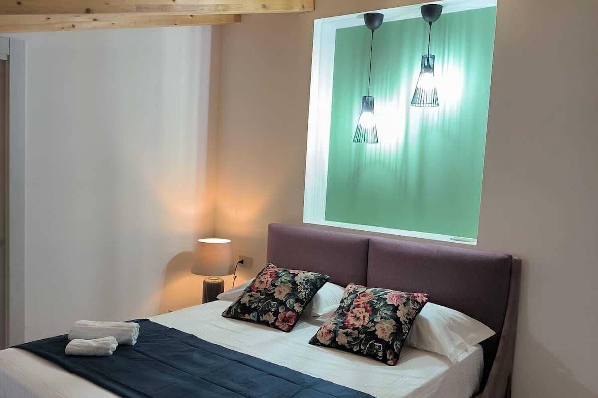 Un dormitorio con cama, almohadas, lámpara de noche, ventana con vistas y vigas de madera en el techo.