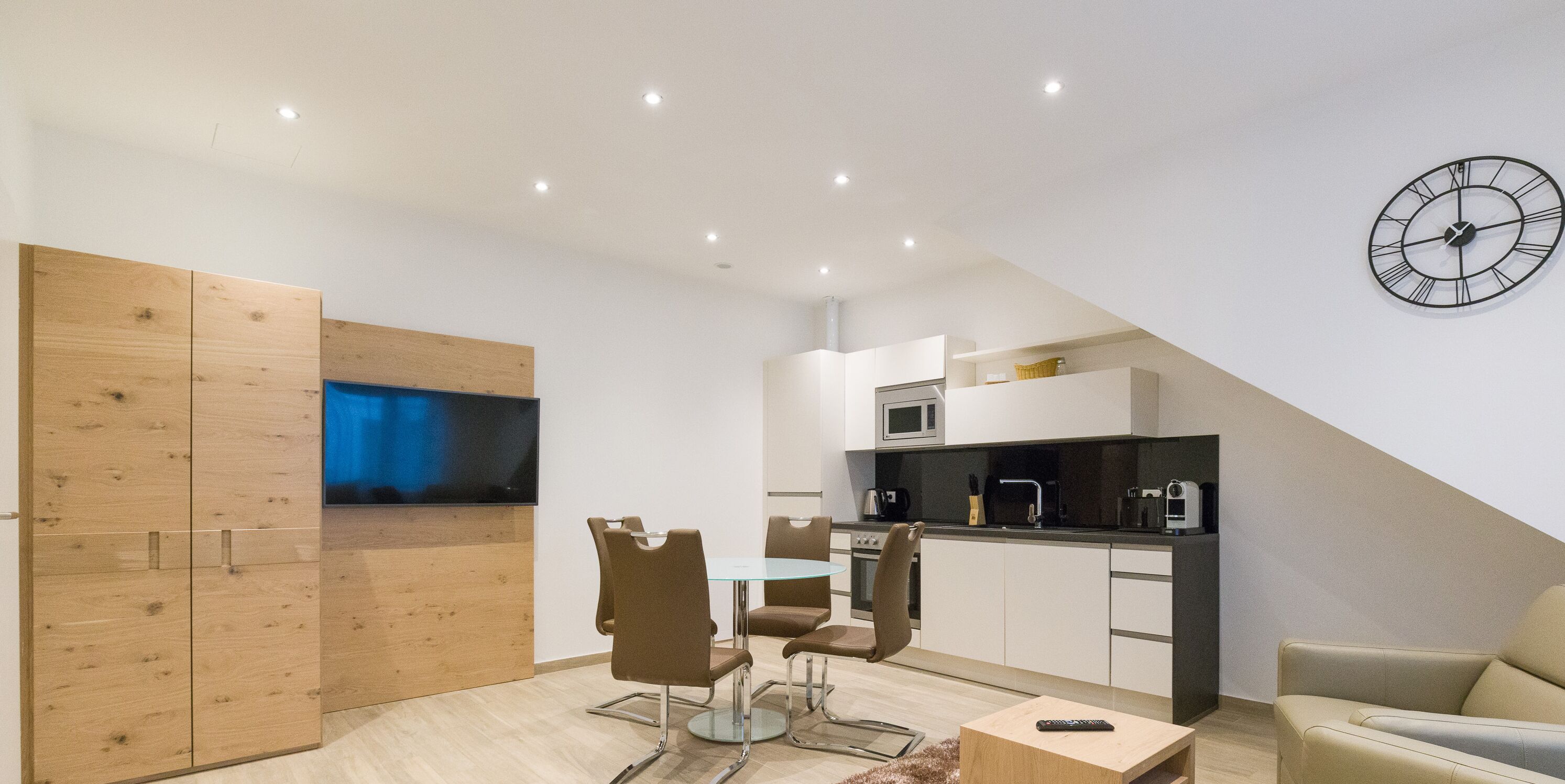 A modern kitchen with a microwave, stove, and coffee maker. There is a dining area with a table and chairs, and a TV mounted on a wooden cabinet.