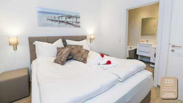 A neatly made bed with white linens and a single red rose, a wall-mounted painting of a beach scene, and a bathroom visible through an open door. - Stadtwohnung mit Flair 1200 (Vienna)