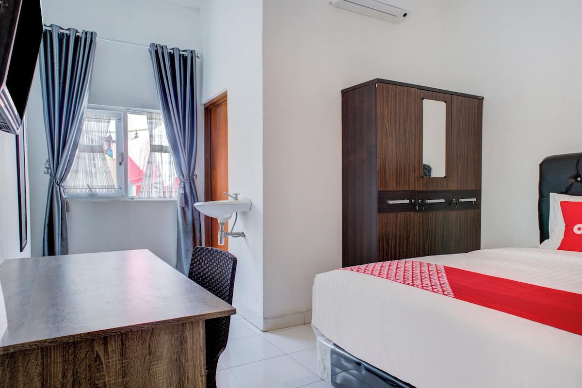 A hotel room with a wooden desk, a bed with a red accent, a wardrobe with a mirror, and a window with curtains.